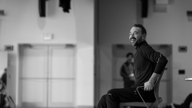 Black-and-white photo of a smiling Kenneth Tindall in a dark long-sleeved top, seated on a chair in a bright rehearsal studio.