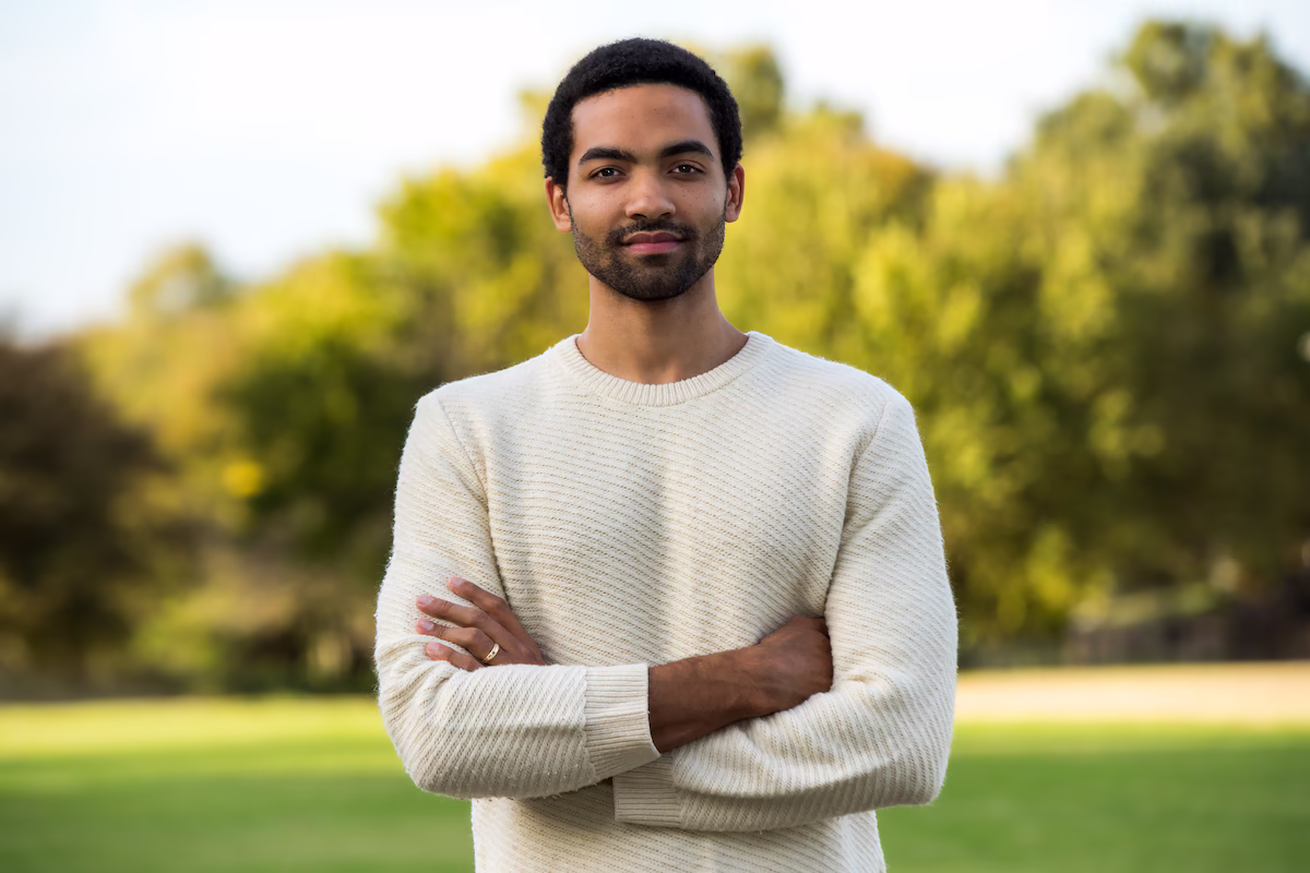 Silas Farley, a former New York City Ballet dancer and award-winning choreographer wearing a cream ribbed sweater, standing with arms crossed in a park with softly blurred green trees behind him.